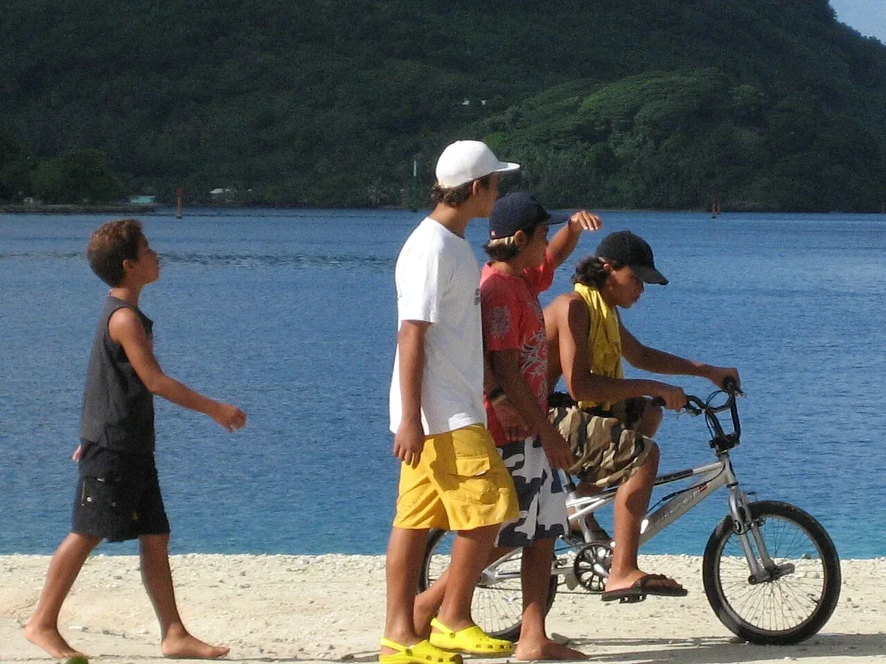 Quatre jeunes marchent et circulent à vélo sur le front de mer de Fare, avec la baie et une colline verdoyante à l’arrière-plan.