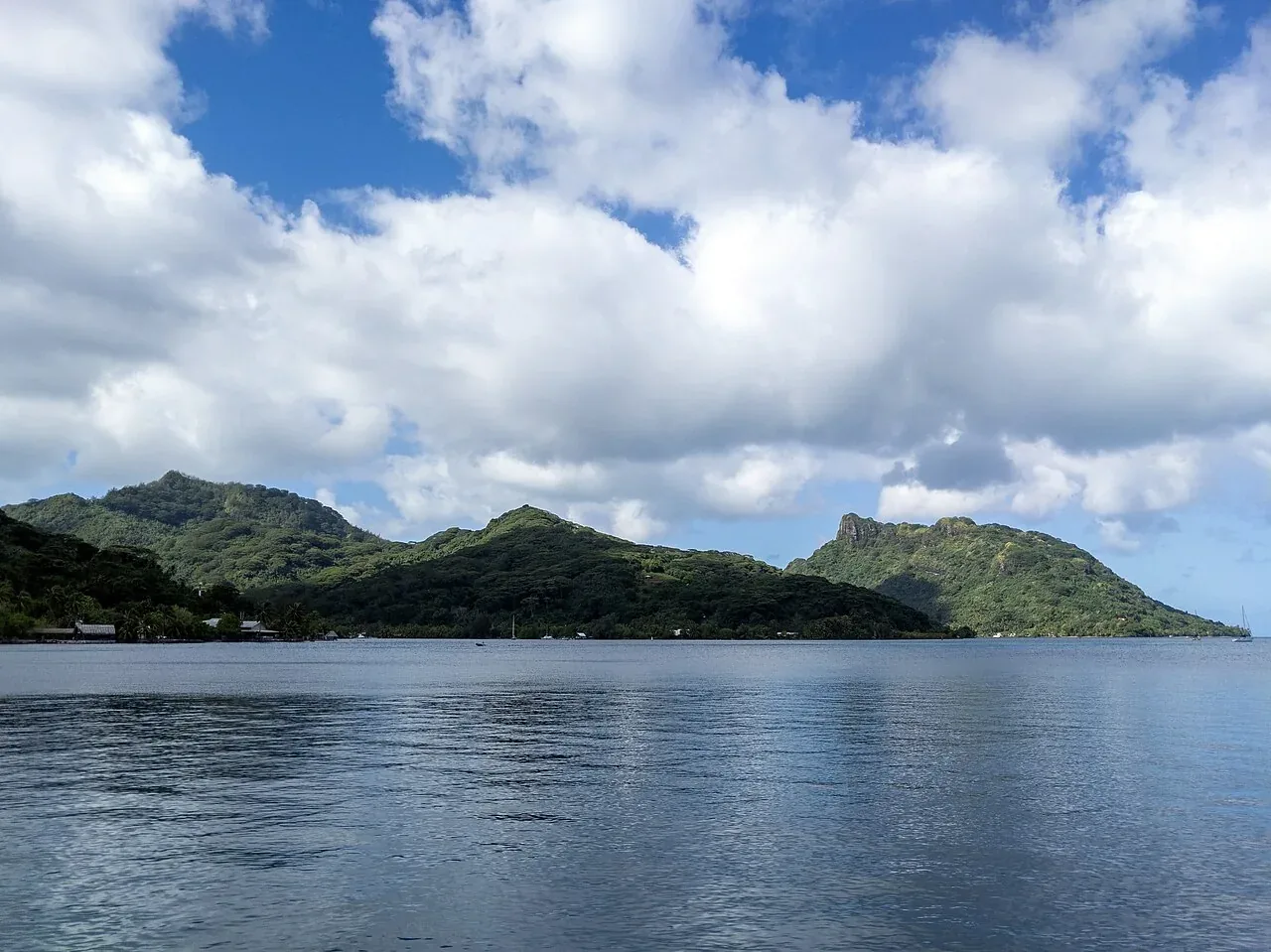 Des collines verdoyantes et des bâtiments du littoral de Huahine Nui, vus au-delà d’une eau calme sous des nuages blancs.