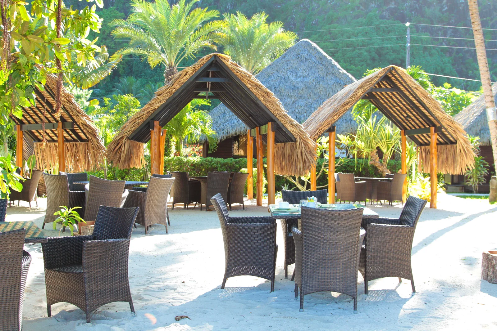 Tables et fauteuils installés sur le sable sous des abris à toiture végétale au restaurant du Mahana.