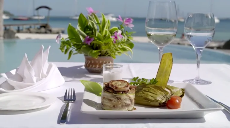 Un plat dressé sur une table du restaurant Omai, avec deux verres, des fleurs et le lagon en arrière-plan.